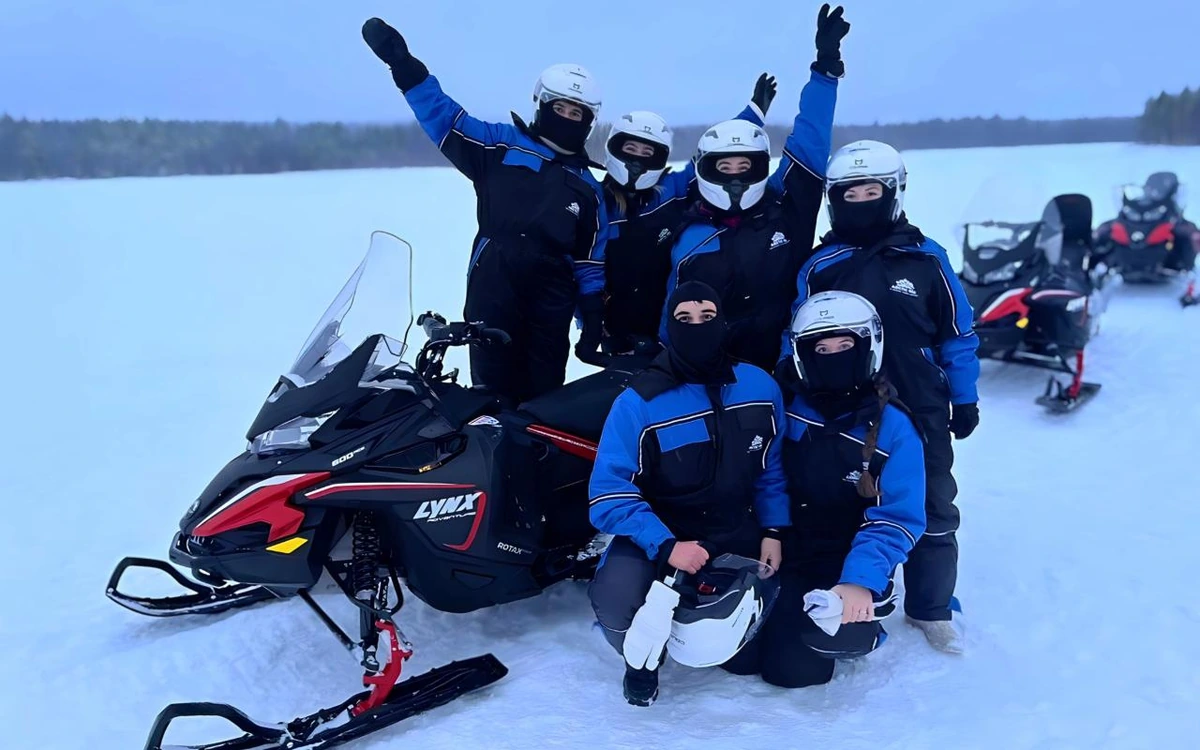 Six riders in snowsuits and helmets beside their snowmobiles on an open snowfield at dusk