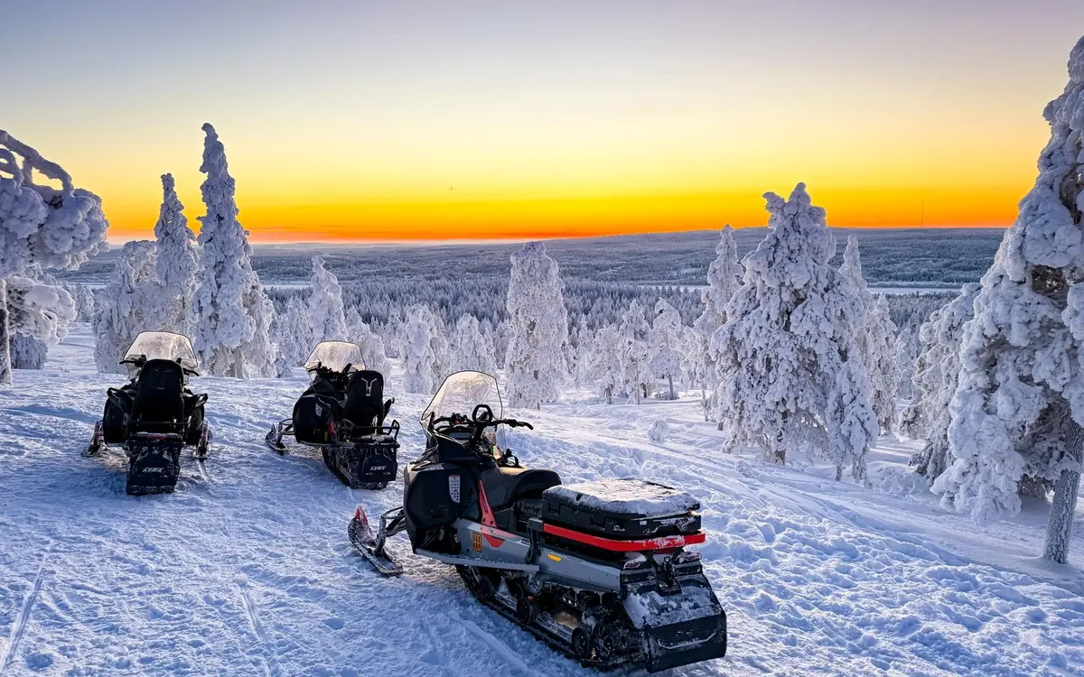 Trois motoneiges à l&rsquo;arrêt sur une crête d&rsquo;arbres givrés sous un coucher de soleil orangé
