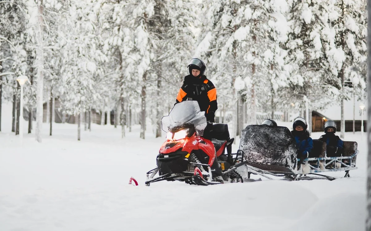 A snowmobile towing a passenger sled with two seated riders through snow-covered pines