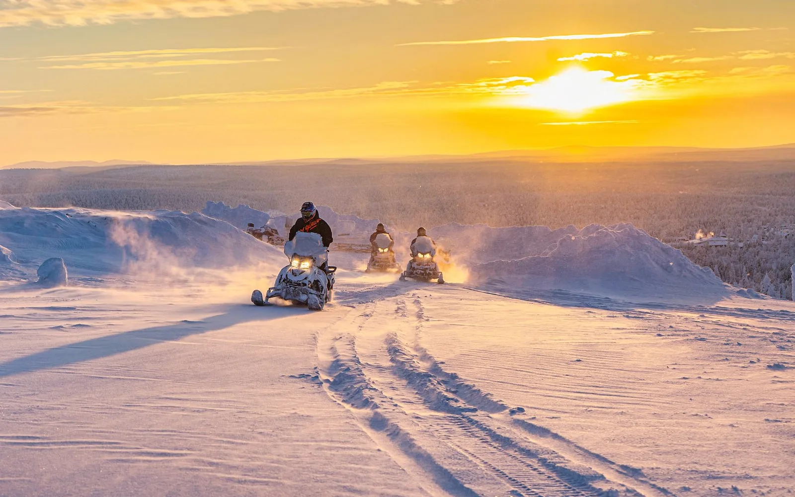 Motoneiges sur une piste damée à travers la neige à perte de vue, soleil bas sur la forêt