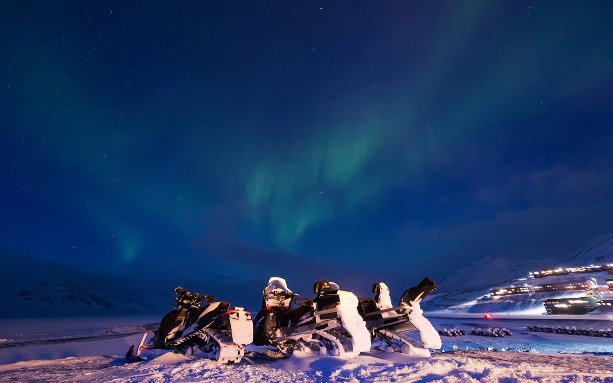 Snowmobiles parked on a snowy ridge under a green and blue aurora